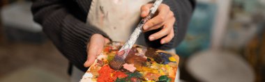 Cropped view of woman in apron holding palette and paintbrush, banner 