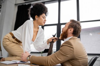 side view of passionate african american woman sitting on desk and pulling tie of bearded coworker in office 