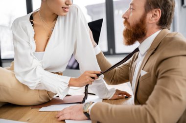 african american businesswoman sitting on desk and pulling tie of tattooed man in office 