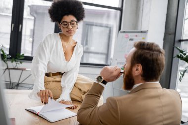 african american woman in glasses sitting on desk and turning page on notebook while looking at bearded coworker  clipart