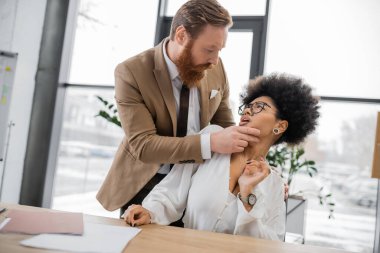 bearded businessman touching face of shocked african american woman in office 