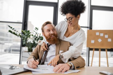 seductive african american woman leaning on shoulders of cheerful man while pointing at charts 