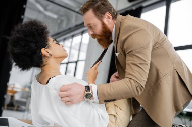 side view of curly african american woman pulling tie of bearded coworker while lying on office table