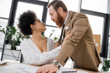 smiling african american woman pulling tie of passionate businessman in office 