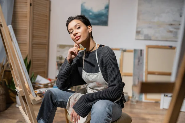 stock image Brunette artist holding paintbrush and looking at camera near blurred canvases in workshop 