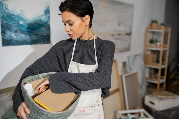 stock image Short haired artist in apron holding basket with equipment in studio 