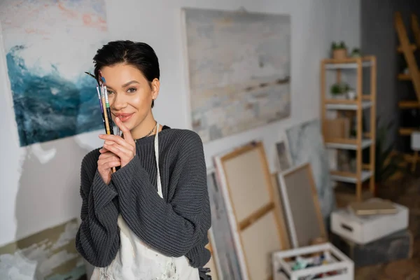 stock image Smiling artist in apron holding paintbrushes near face in studio 