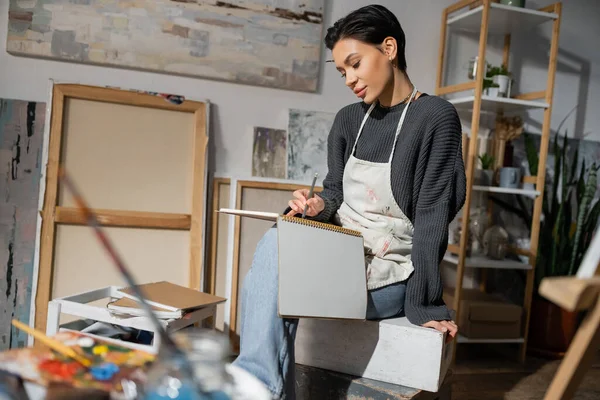 stock image Young artist drawing on sketchbook while sitting near paintings in studio 