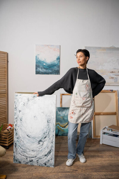 Короткие волосы художника, стоящего с рукой на бедре возле живописи в мастерской 