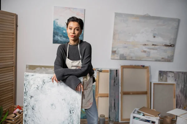 stock image Young artist looking at camera while standing near painting in studio 