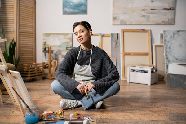 Young artist in apron holding paintbrush near paints and canvas in workshop 