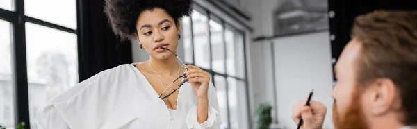 stock image sexy african american businesswoman holding glasses and looking at blurred coworker, banner 