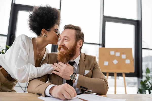 Stock image smiling african american businesswoman hugging while whispering in ear of cheerful coworker in office 