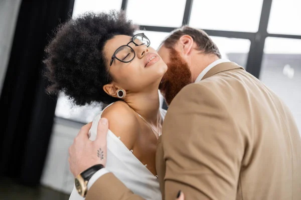 tattooed man undressing and kissing neck of seductive african american woman in office