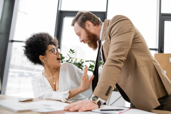 shocked african american woman screaming and pushing away businessman in office 