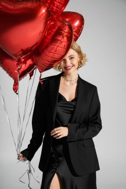 happy young woman in black slip dress and blazer holding red heart-shaped balloons isolated on grey 