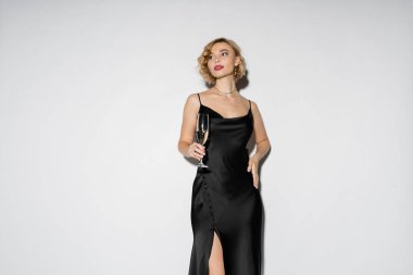 blonde woman in satin slip dress holding glass of champagne while standing with hand on hip isolated on grey 