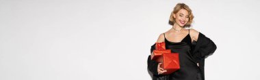 happy woman in black blazer and satin slip dress holding red presents while posing with hand on hip on grey, banner 