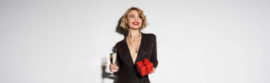 happy young woman in tight black dress holding red gift box and glass of champagne on grey, banner 