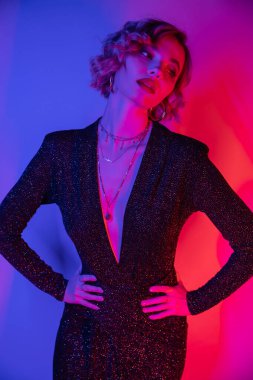 young woman with blonde hair posing with hands on hips in black tight dress on purple and pink 