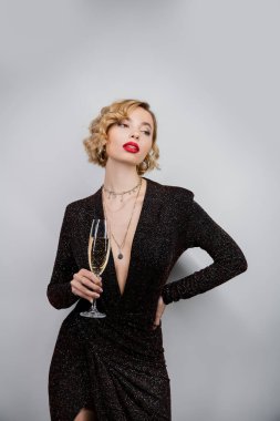 young woman with blonde curly hair and red lips posing with hand on hip while holding glass of champagne on grey 