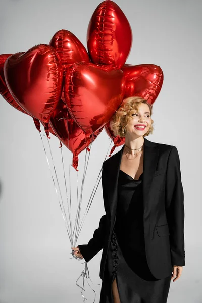 cheerful woman in black slip dress and blazer holding red heart-shaped balloons on grey