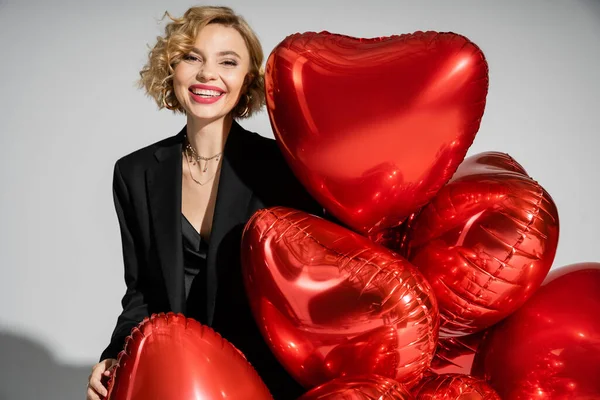 happy young woman in black slip dress and blazer smiling near red heart-shaped balloons on grey