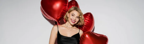 cheerful young woman in black and satin slip dress posing near red balloons on valentines day on grey, banner