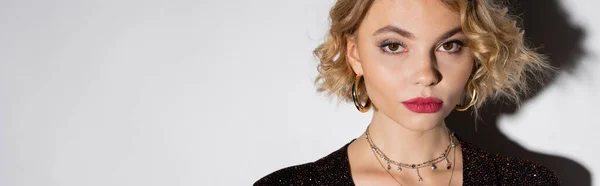 stock image portrait of blonde young woman in golden hoop earrings and silver necklace looking at camera on white, banner 