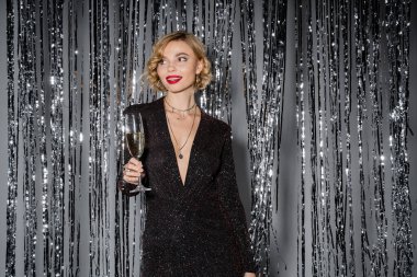 cheerful young woman in black tight dress standing with glass of champagne near shiny tinsel curtain on grey