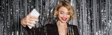 cheerful woman taking selfie while holding glass of champagne near tinsel curtain on grey, banner 