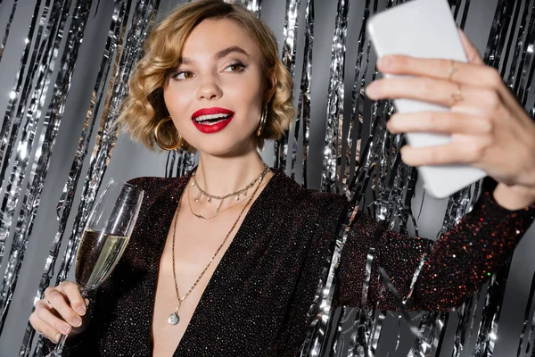 stock image cheerful woman in black dress taking selfie while holding glass of champagne near tinsel curtain on grey