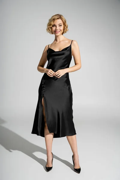full length of cheerful and blonde woman in elegant slip dress standing on grey