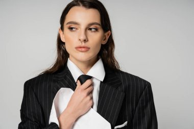 brunette woman in elegant oversize outfit adjusting tie and looking away isolated on grey