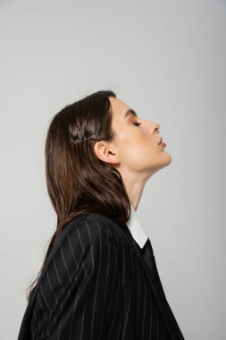 side view of young brunette woman in black and striped blazer isolated on grey