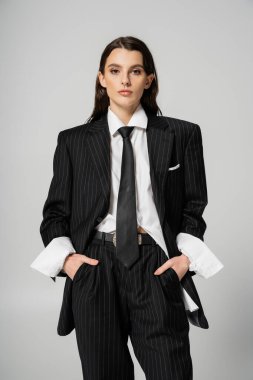 front view of brunette woman in stylish oversize suit holding hands in pockets while looking at camera isolated on grey