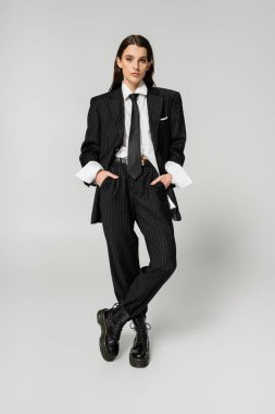 full length of brunette woman in elegant oversize pantsuit and rough boots standing with hands in pockets on grey background