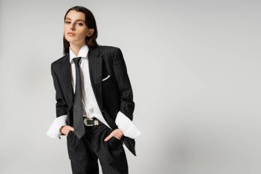 pretty woman in black suit and white shirt standing with hands in pockets while looking at camera isolated on grey