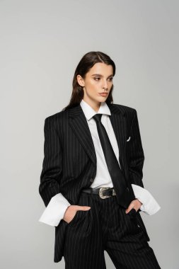 young brunette woman in black suit and white shirt standing with hands in pockets isolated on grey