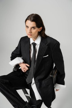 brunette woman looking at camera while sitting in black stylish pantsuit isolated on grey