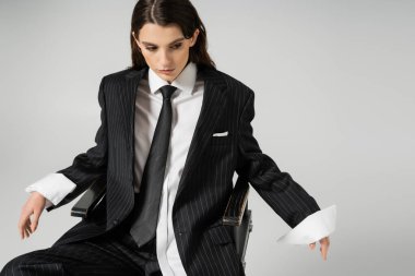 young woman in white oversize shirt and black striped suit sitting on chair isolated on grey