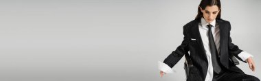 young woman in white shirt and black blazer with tie looking down while sitting isolated on grey, banner