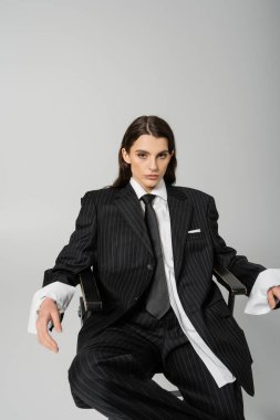brunette model in black striped suit and tie sitting on chair and looking at camera isolated on grey