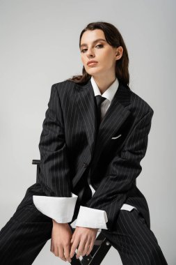 elegant woman in black oversize suit sitting and looking at camera isolated on grey