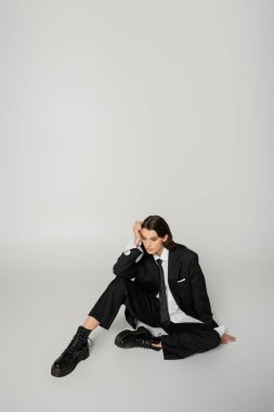 pretty woman in laced-up boots and oversize formal wear sitting with hand near head on grey background