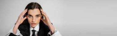 portrait of brunette woman in stylish attire touching head while looking at camera isolated on grey, banner