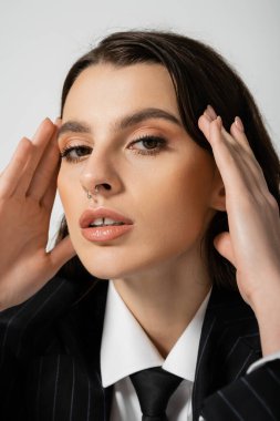 portrait of fashionable woman with makeup and piercing looking at camera and holding hands near face isolated on grey