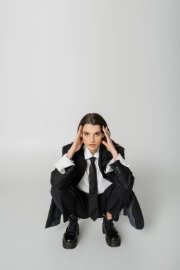 brunette woman in black striped suit and rough boots holding hands near face and looking at camera while sitting on grey 