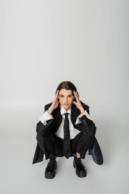 brunette woman in black striped suit and rough boots holding hands near face and looking at camera while sitting on grey  clipart