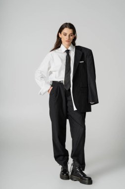 full length of young model in oversize formal wear and rough boots standing with hand in pocket on grey background